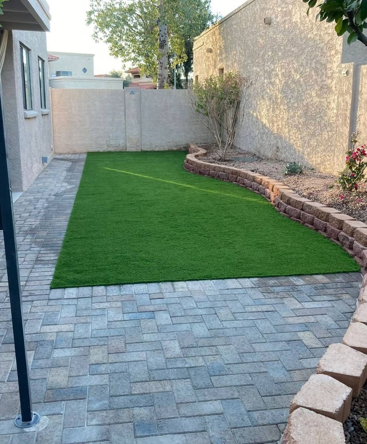 Finished backyard by Little Santi: artificial turf bordered by pavers and a curved block planter wall