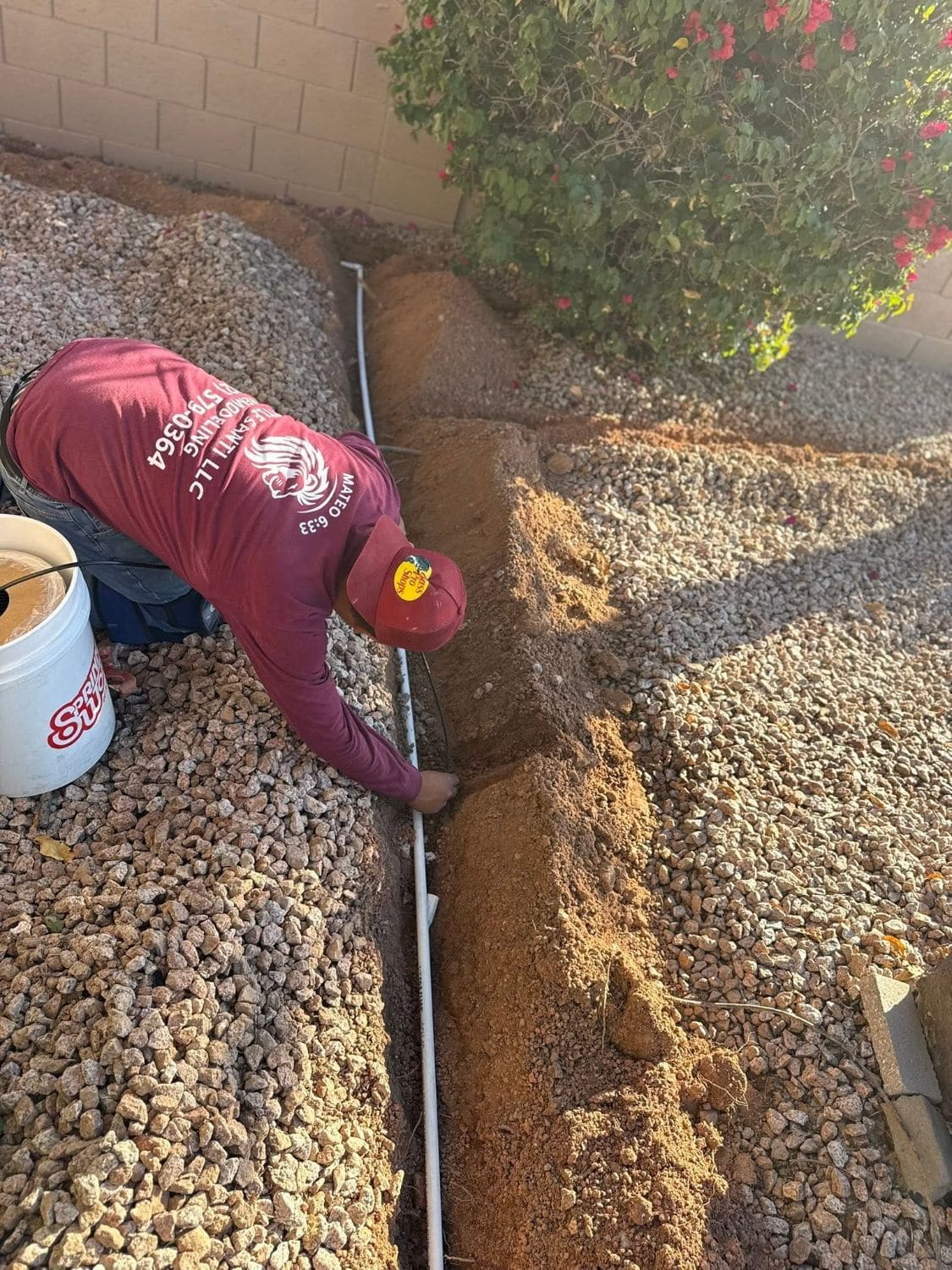 A Little Santi crew member in a branded shirt laying irrigation pipe in a trench through a rock bed