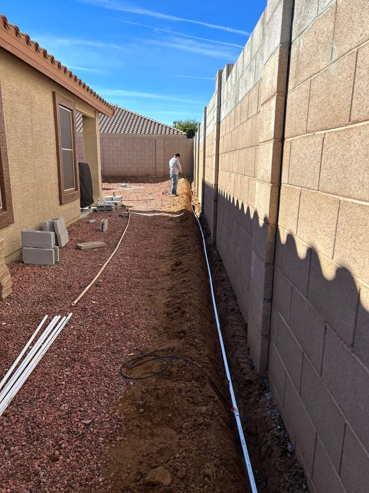 Long trench cut along a block wall through a red-rock side yard, with pipe laid in