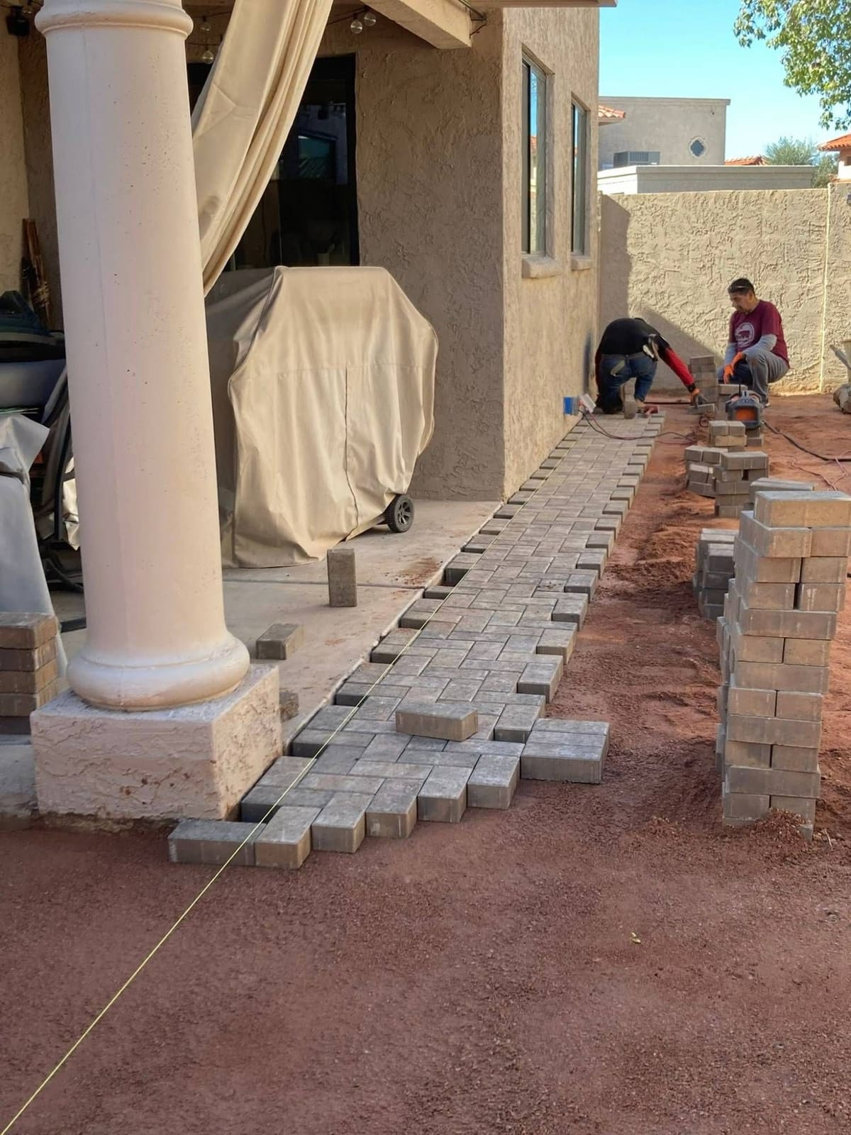 Crew laying rectangular pavers along a covered patio, with stacked pavers ready nearby